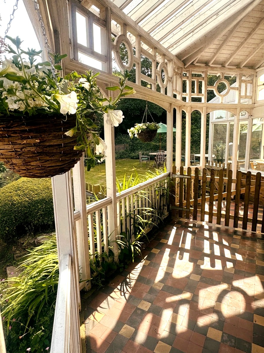 Inside the white-painted Victorian conservatory at St Anselm's, with hanging baskets of white petunias, decorative fretwork, a pitched glass roof, checkerboard tiled floor and the garden visible through the railings